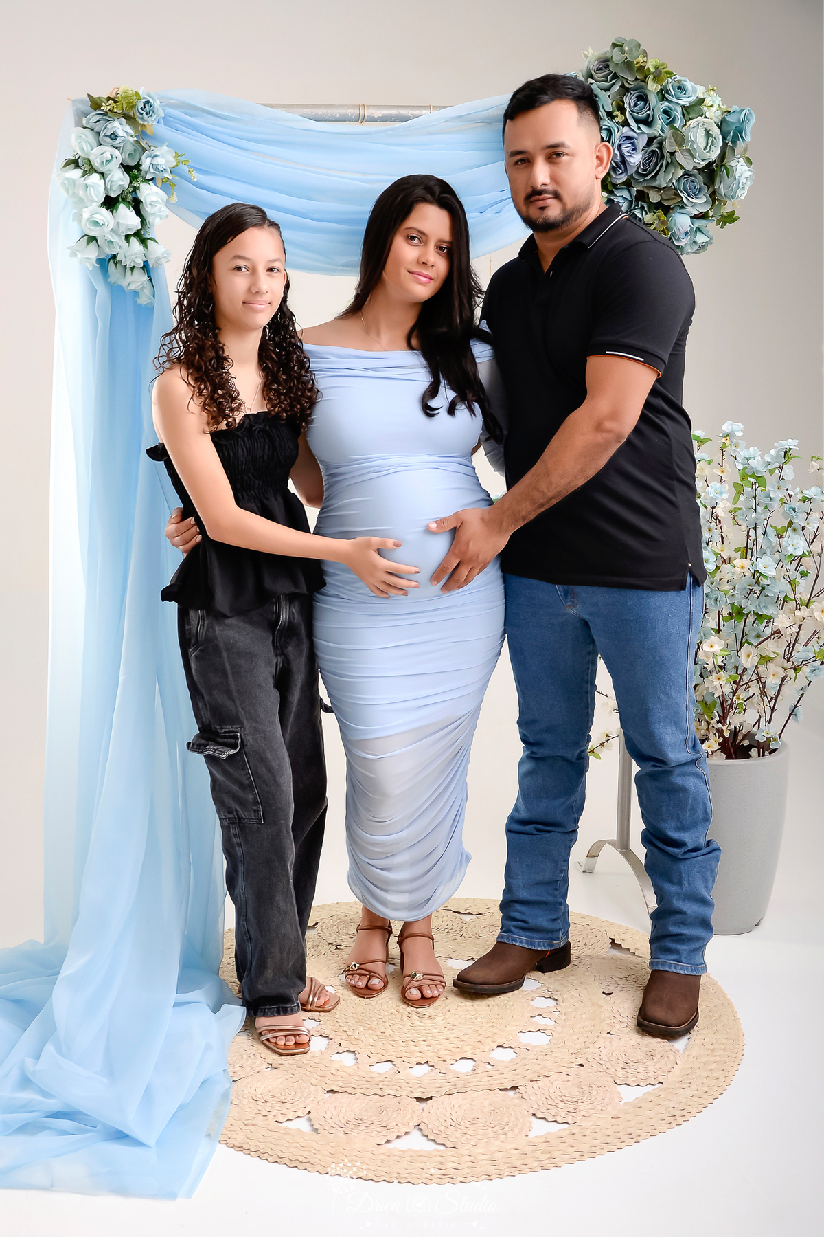 drica studio - xinguara - pará- familia - mãe , pai e filhos - mamãe de segunda viagem -  mamãe  usando vestido azul - pai d filha usando calça jeans e camisa preta - flores ao fundo 