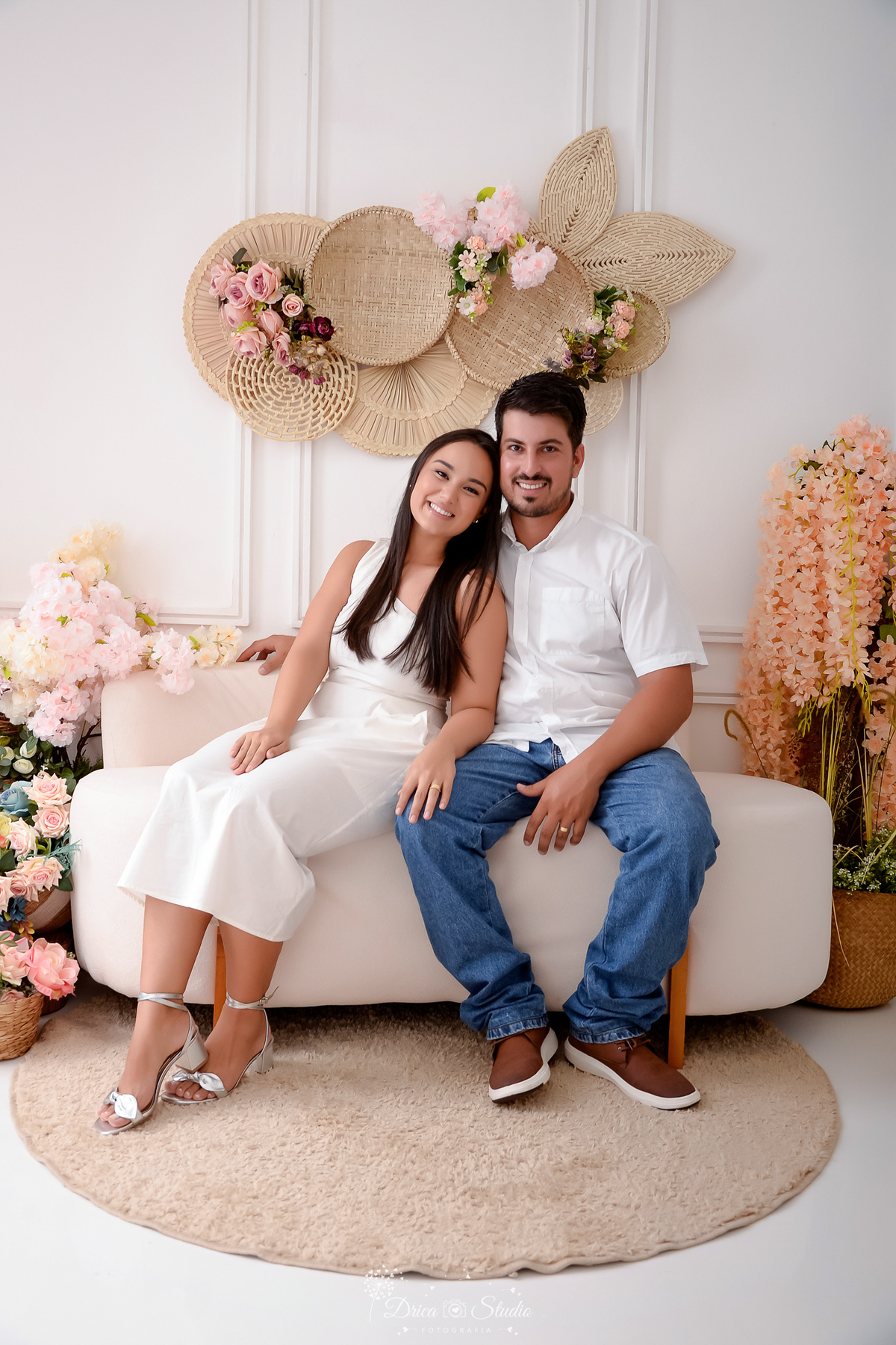 drica studio - fotos familia - xinguara - pará - casal - sentados em um sofá - cenário com flores ao fundo - casal usando roupas brancas 