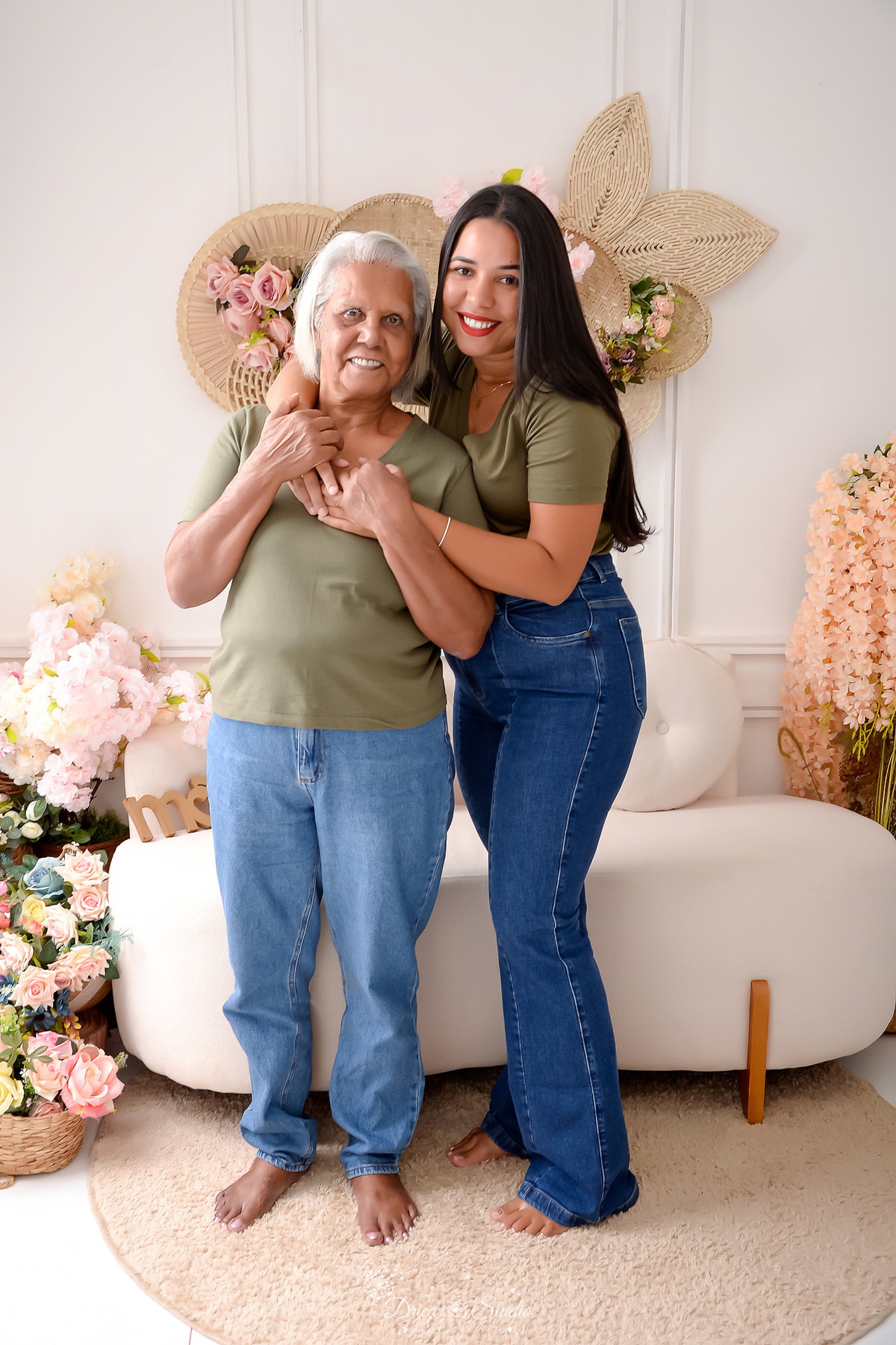 drica studio - ensaio familia - 4 gerações - bizavó - avó - mãe - filha - netá e bizavó abraçadas -  em cenário com fundo branco e flores ao fundo - usando camisa verde no mesmo tom - e calça jeans - familia reunida 