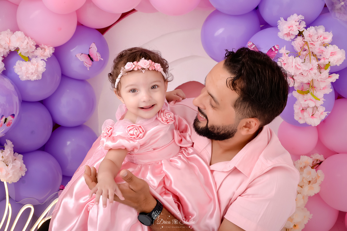 drica studio - smash the cake - jardim encantado - fotos em estudio - pai e filha - cenário com balões rosas e lilás - flores e borboletas - bebê usando vestido rosa - pai usando camisa rosa 