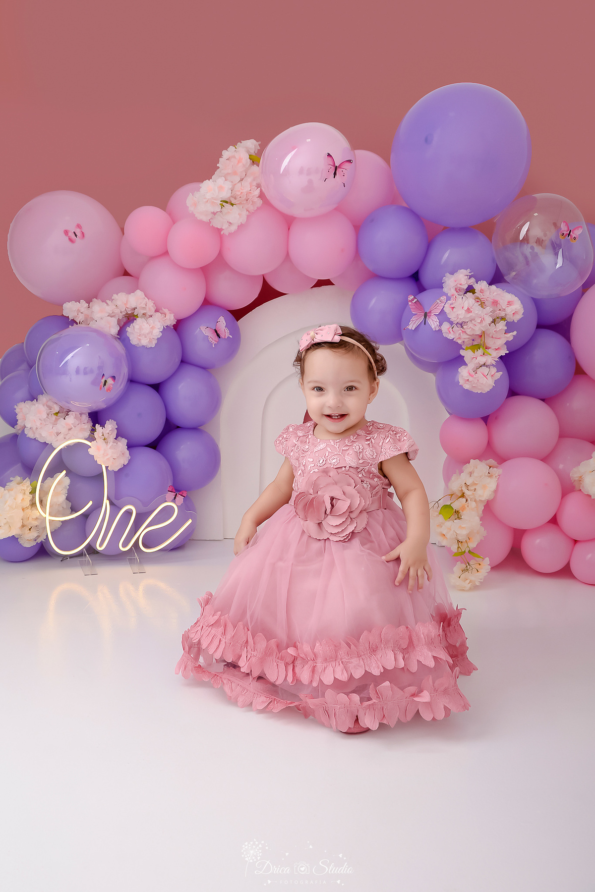 drica studio - smash the cake - jardim encantado - fotos em estudio - cenário com balões rosas e lilás - com flores e borboletas - one de led - bebê usando vestido rosa - sentanda num banquinho 