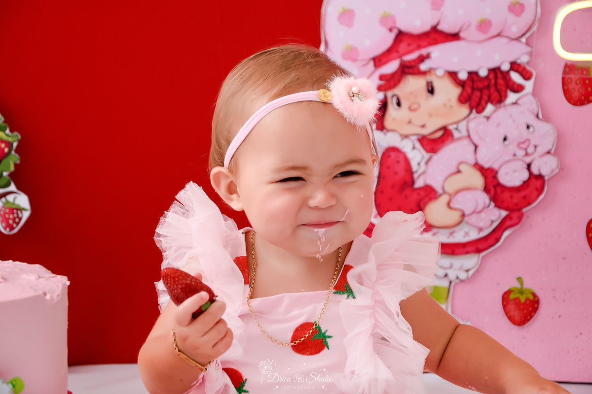 dricastudio - smash the cake - acompanhamento mensal - 1 aninho - xinguara - pará -bebê comendo um morango - ao fundo a imagem da moranguinho - bebê usando vestido e laço 