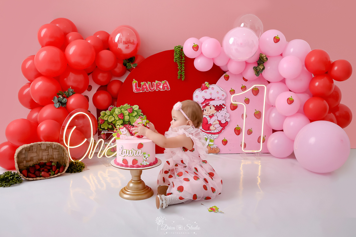 dricastudio - smash the cake - acompanhamento mensal - 1 aninho - xinguara - pará - cenário com balões rosas e vermelhos - monrangos pelo cenário - one de led - 1 de led - primeiro contato com o bolo - bebê usando vestido de morangos - usando laço 