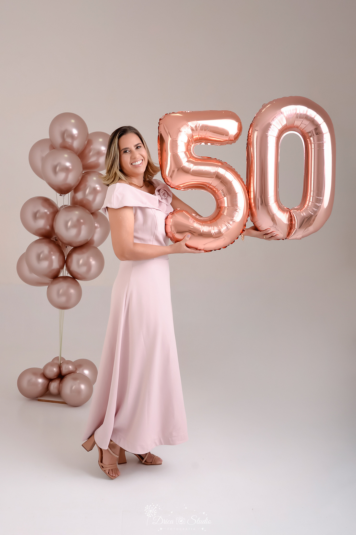 Ensaio comemorativo de 50 anos realizado em estúdio com iluminação suave e elegante.
Cliente vestindo um sofisticado vestido em tom rosa claro, transmitindo delicadeza e feminilidade.
Sorriso espontâneo e acolhedor, 