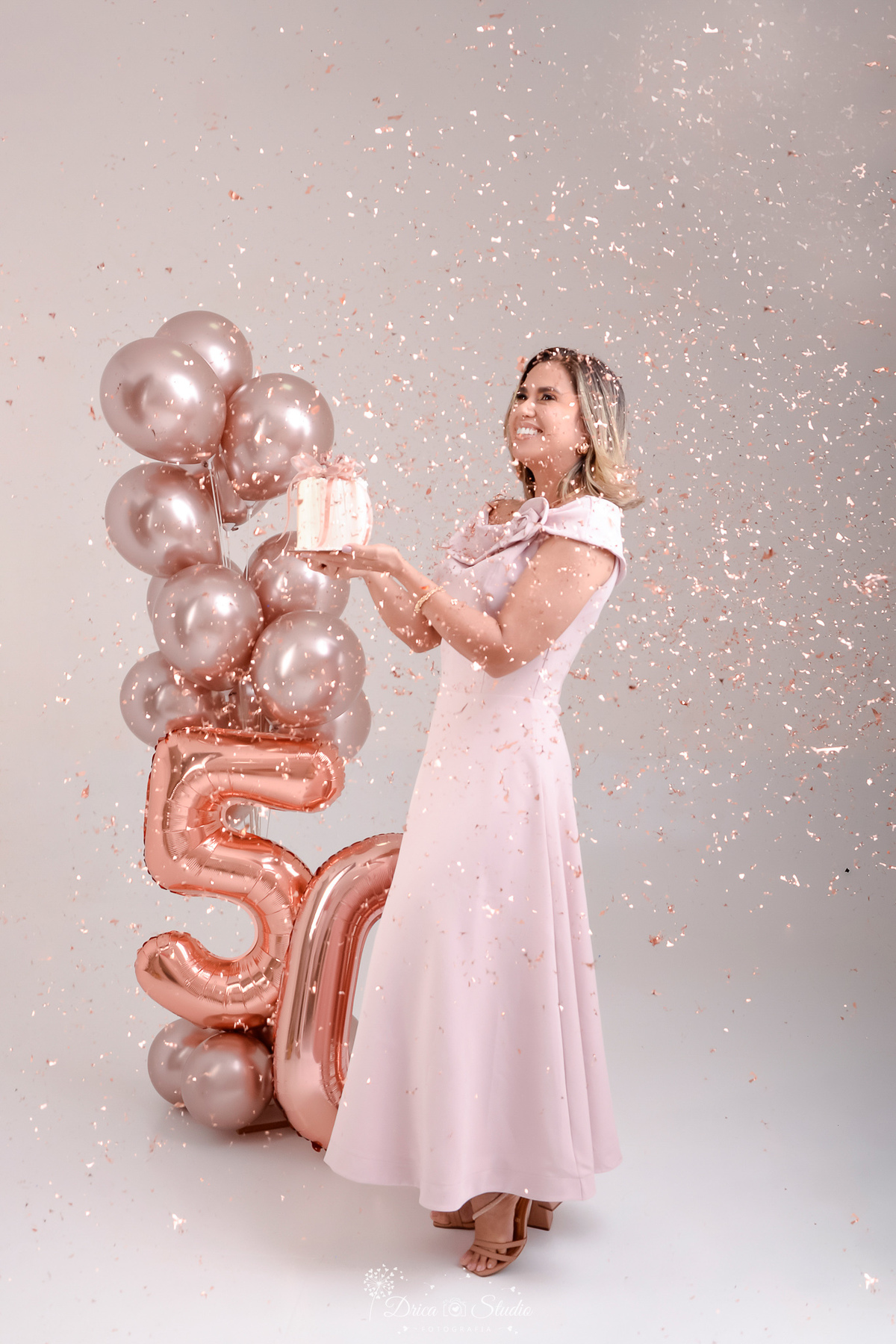 Ensaio comemorativo de 50 anos realizado em estúdio com iluminação suave e elegante.
Cliente vestindo um sofisticado vestido em tom rosa claro, transmitindo delicadeza e feminilidade.
Sorriso espontâneo e acolhedor, 