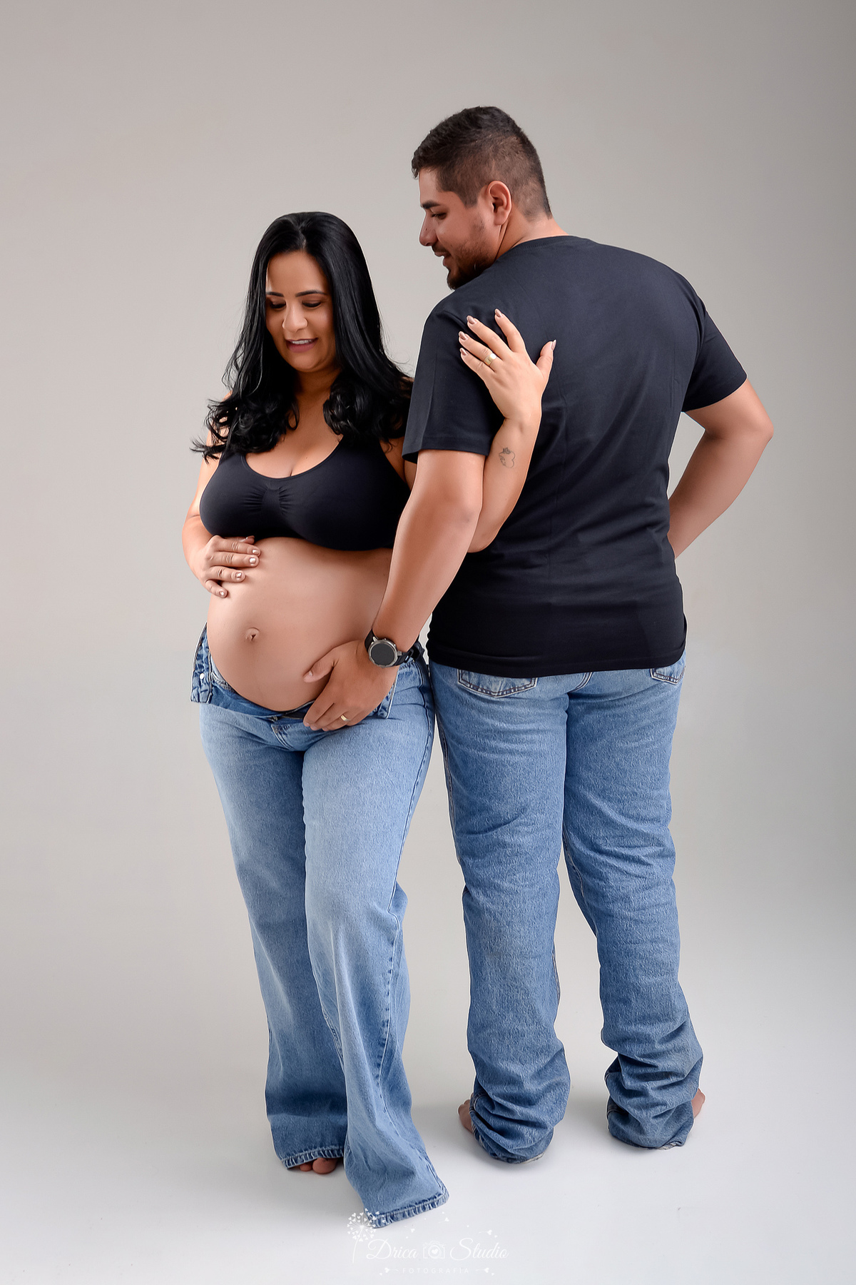 EnsaioGestante - MeninaACaminho - ÀEsperaDaPrincesa - PrimeiraFilha - FotografiaGestante Família - Gestação - MemóriasParaSempre - drica studio - fotos em studio - gestante usando calça jeans e tope preto - esposo usando camisa preta e calça jeans 