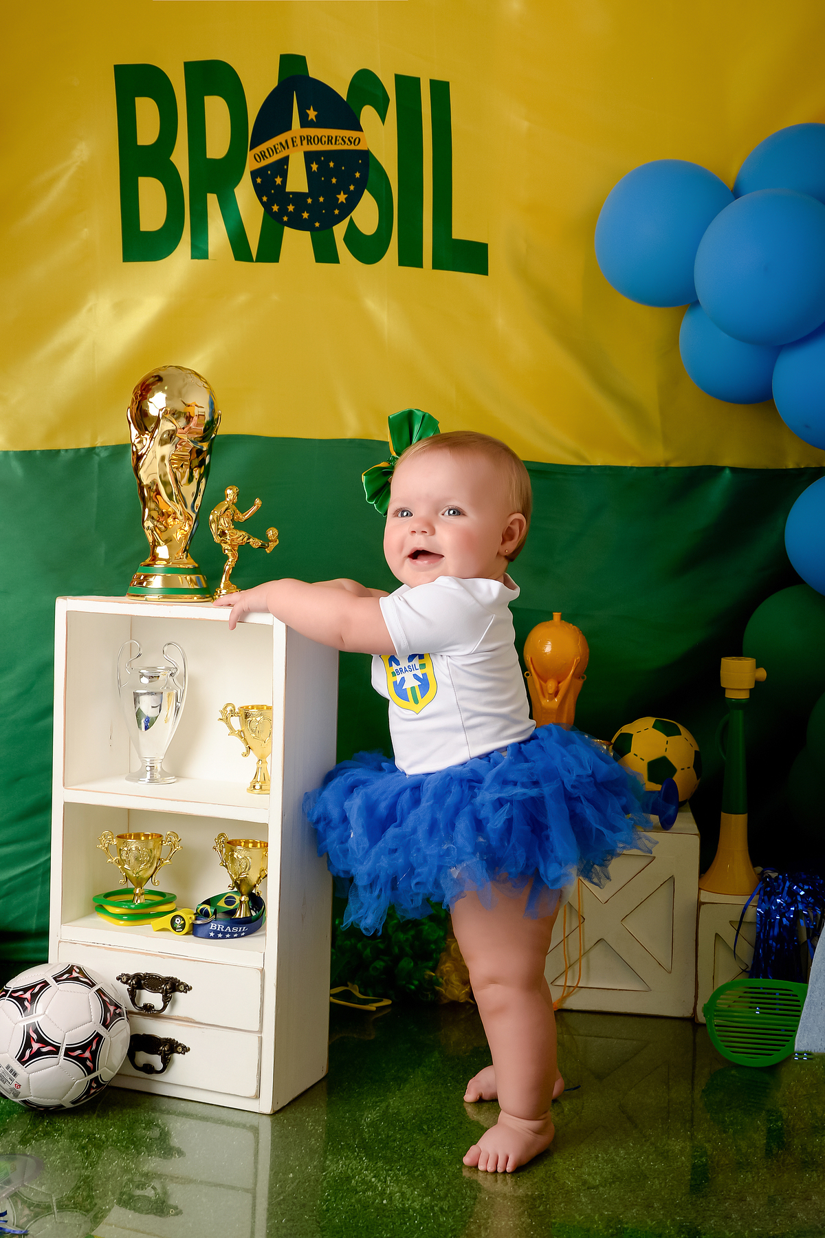 Drica Studio - Copa2026 -Lembrança especial primeira copa - cenário de copa - verde, amarelo e azul - bebê usando bory branco e saia de tulê azul - trofeu da copa - bolas e trofeis no cenário 