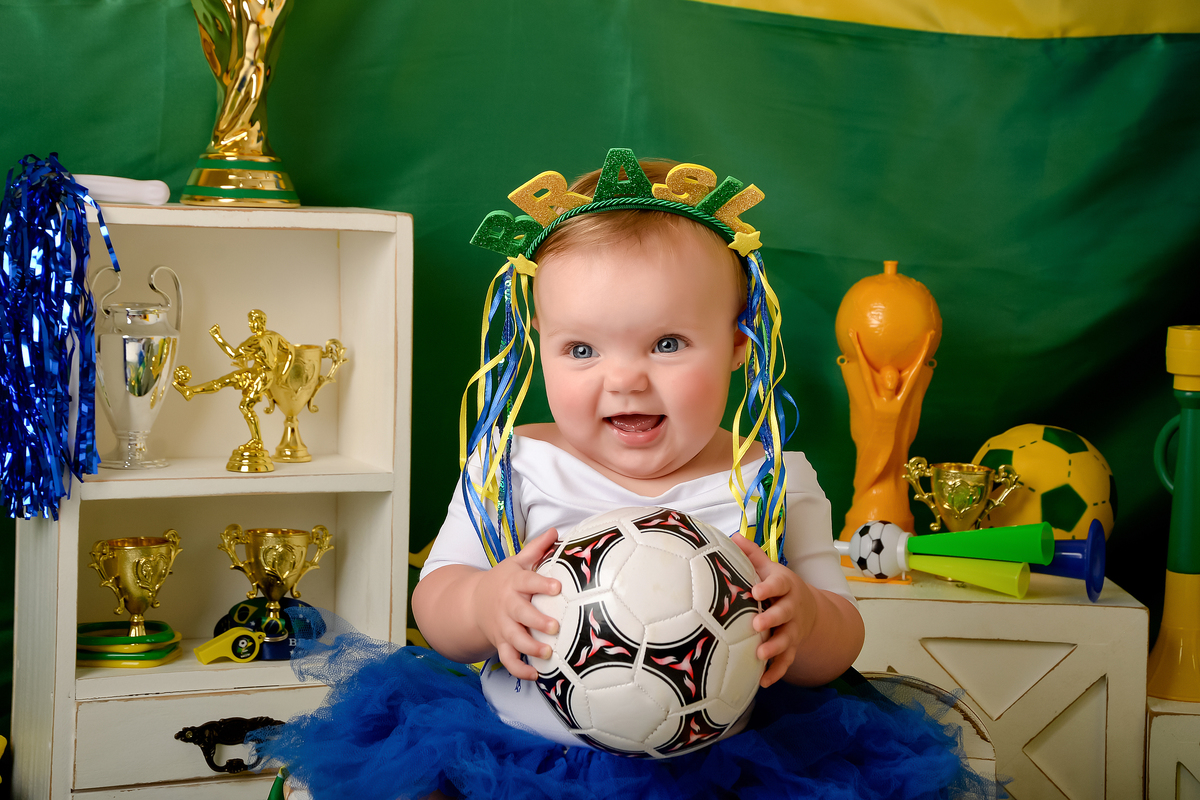 Drica Studio - Copa2026 -Lembrança especial primeira copa - cenário de copa - verde, amarelo e azul - bebê usando bory branco do brasil e saia de tule azul - com tiara do brasil - segurando uma bolo de futbol 