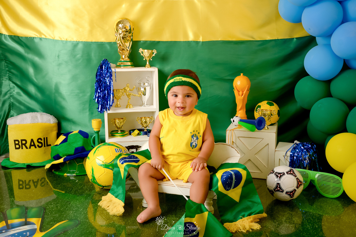 Drica Studio - Copa2026 -Lembrança especial primeira copa - cenário de copa - verde, amarelo e azul -bebê usando bory amarelo do brasil - segurando bandeira - sentado em um banquinho - bolas de futbol no cenário - bebê usando faixa do brasil 