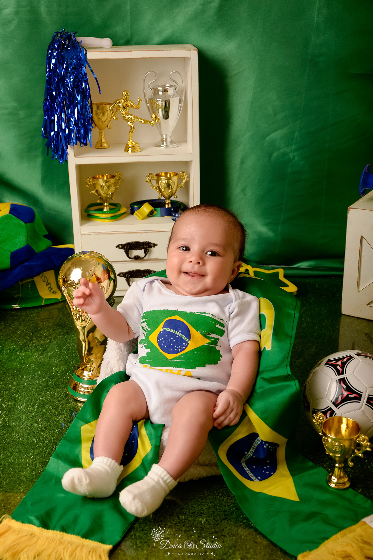 Drica Studio - Copa2026 -Lembrança especial primeira copa - cenário de copa - verde, amarelo e azul - bebê usando bory branco do Brasil - com taça da cops ao lado - sorrindo 