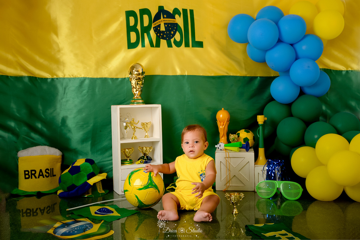Drica Studio - Copa2026 -Lembrança especial primeira copa - cenário de copa - verde, amarelo e azul - bebê sentado no chão com bola amarela de futbol - usando bory amarelo - cenário com fundo amarelo e verde - com balões nas cores da badeira 