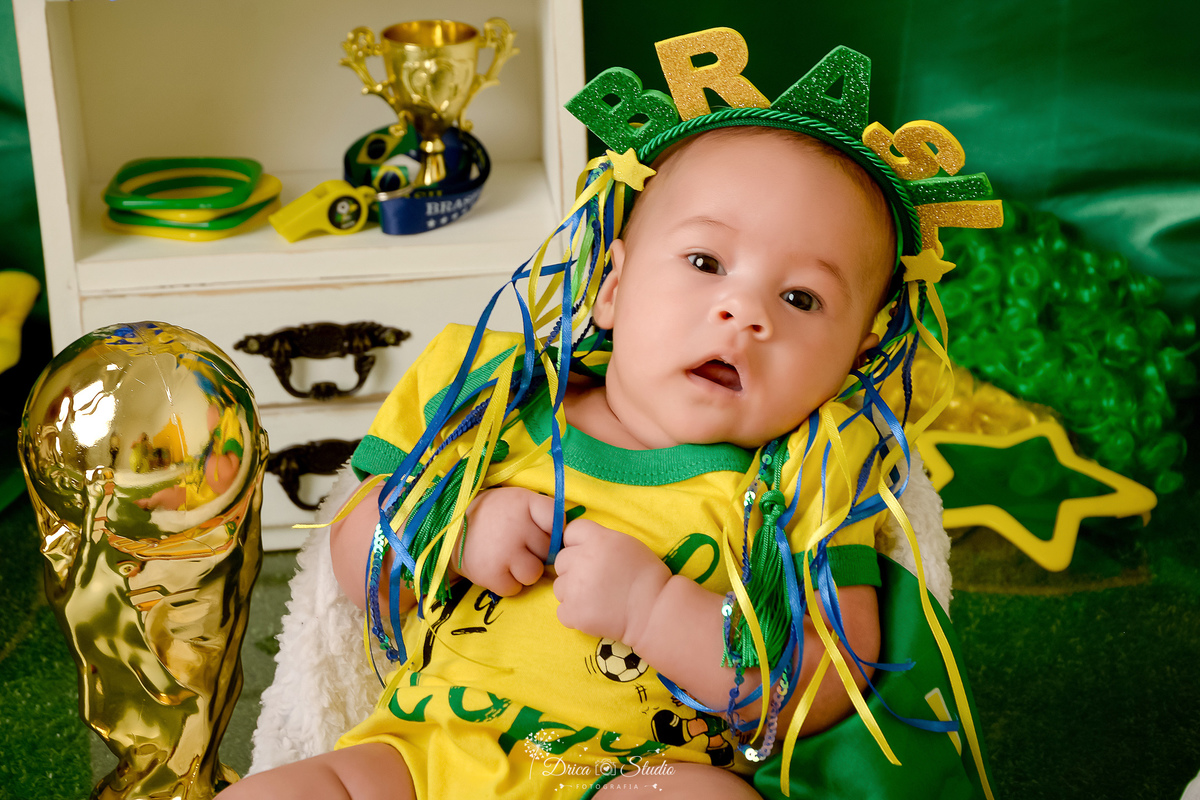 Drica Studio - Copa2026 -Lembrança especial primeira copa - cenário de copa - verde, amarelo e azul - bebê usando bory amarelo - com tiara do brasil - com a taça da copa ao lado - 