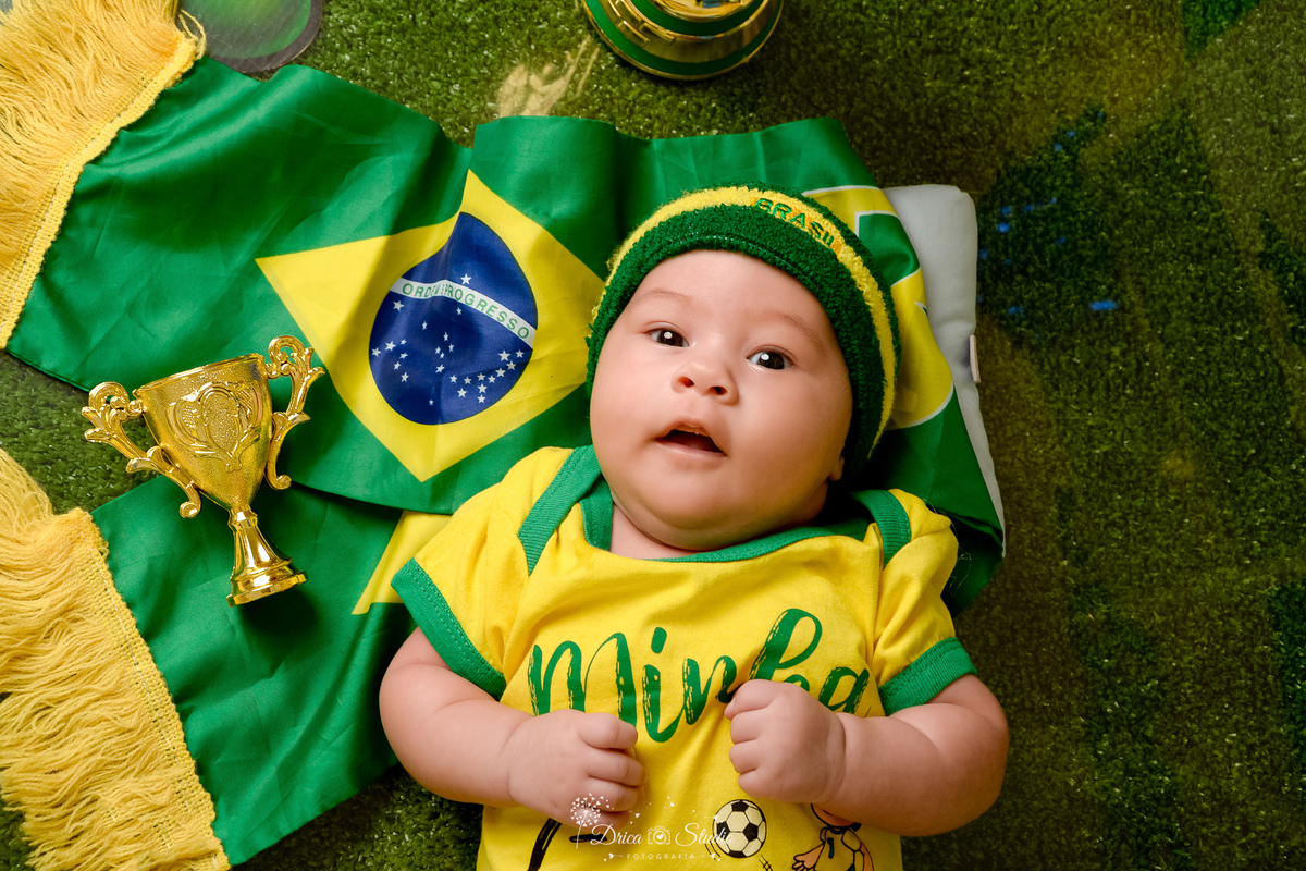 Drica Studio - Copa2026 -Lembrança especial primeira copa - cenário de copa - verde, amarelo e azul - bebê usando bory amarelo de copa - com faixa do brasil na cabeça - deitada sobre a bandeira do brasil 