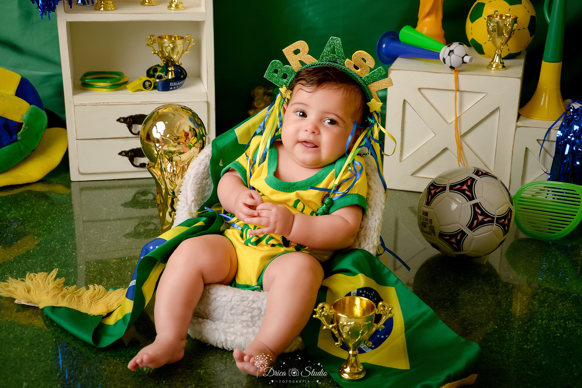 Drica Studio - Copa2026 -Lembrança especial primeira copa - cenário de copa - verde, amarelo e azul - bebê usando bory do brasil - com tiara do brasil - ao lado de uma bola de futbol - sentada em um banquinho branco 