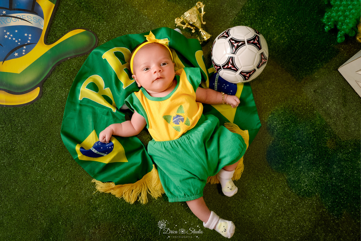 Drica Studio - Copa2026 -Lembrança especial primeira copa - cenário de copa - verde, amarelo e azul - bebê usando bory e shor do brasil - deitada sobre a bandeira do brasil  - com uma bola de futbol do lado 