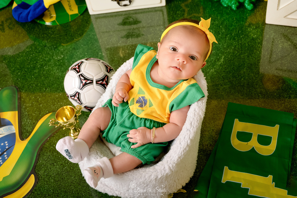 Drica Studio - Copa2026 -Lembrança especial primeira copa - cenário de copa - verde, amarelo e azul - bebê sentada em uma cadeirinha - usando bory e shorts do brasil - com laço amarelo - com uma bola de futbol do lado - 