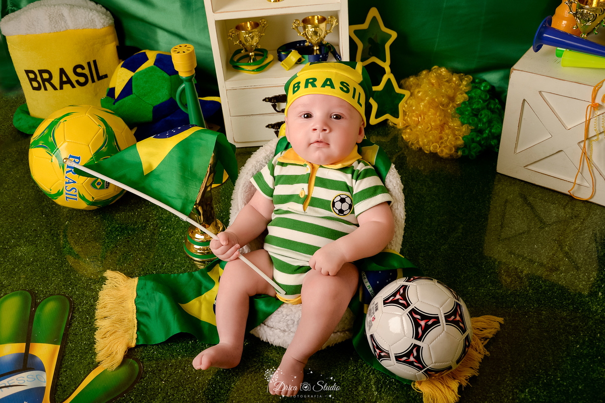 Drica Studio - Copa2026 -Lembrança especial primeira copa - cenário de copa - verde, amarelo e azul - bebê usando bory do brasil - sentado em cadeirinha - segurando uma bandeira do brasil - com uma bola de futbol do lado e com bandana\ na cabeça 