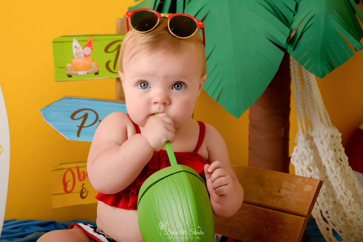 verão2026 - cenário de praia - acompanhamento mesal - drica -studio - xinguara - pará - bebê com copinho de coco - sentada em uma cadeira de praia - com um oculos vermelho na cabeça - fundo amarelo - com um coqueiro verde e marrom ao fundo 