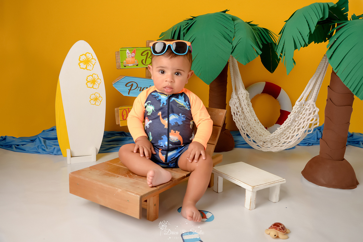 verão2026 - cenário de praia - acompanhamento mesal - drica studio - xinguara - pará - bebê sentado em uma cadeira de praia - ao fundo dois coqueiros verde com marrom e uma rede branca - ao lado uma prancha de surfe 