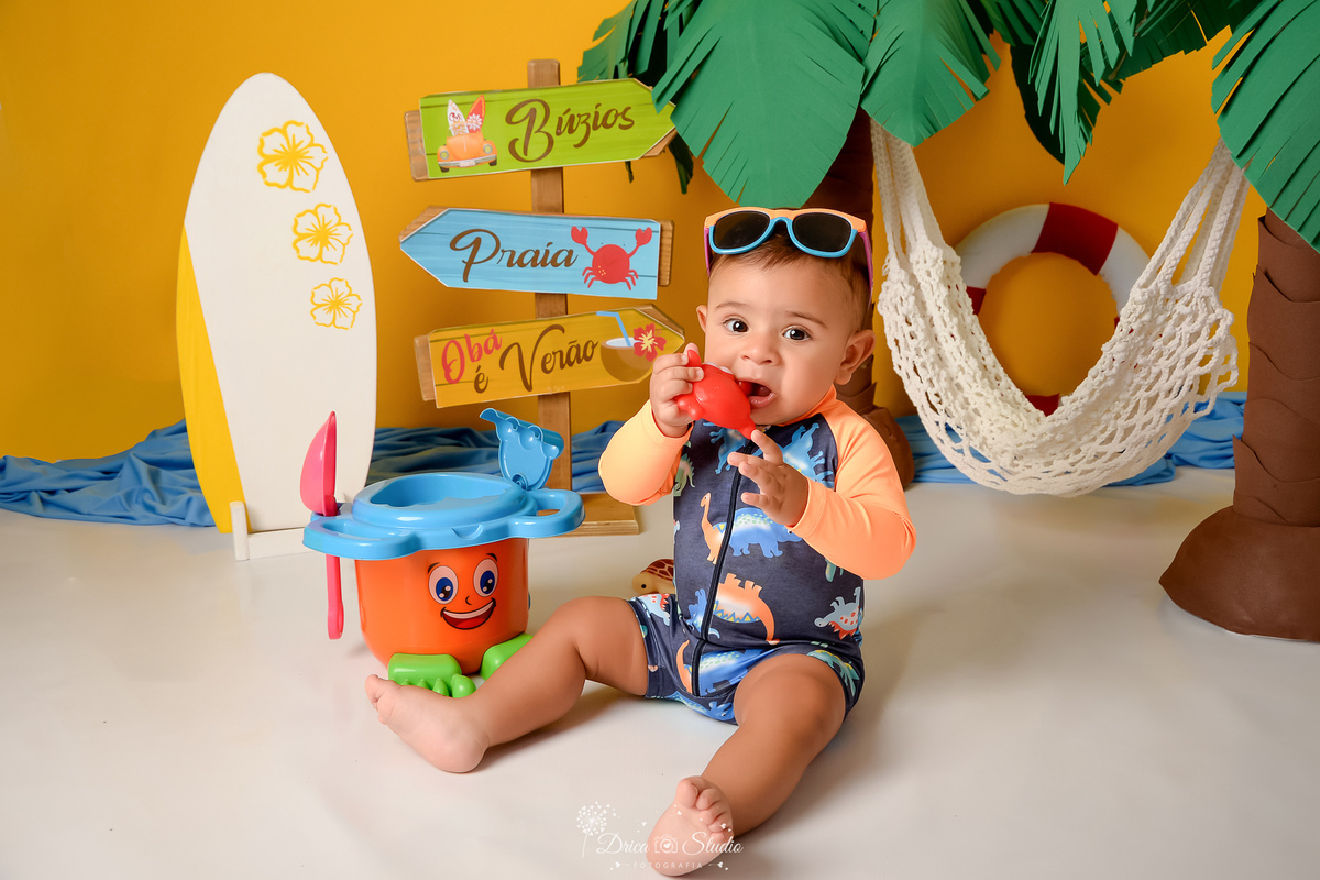 verão2026 - cenário de praia - acompanhamento mesal - drica studio - xinguara - pará - bebê sentado em cenário de praia - fundo amarelo - com dois coqueiros ao fundo - uma prancha de surfe -com um baldinho de areia 