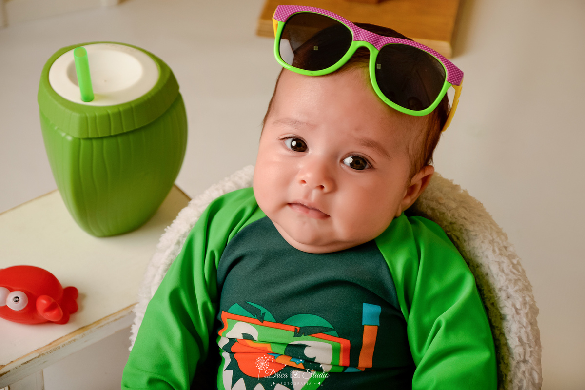 verão2026 - cenário de praia - acompanhamento mesal - drica studio - xinguara - pará - bebê sentado numa cadeirinha branca - com oculos na cabeça - ao lado um copinho em formato de coco - usando rouoa de banho verde 