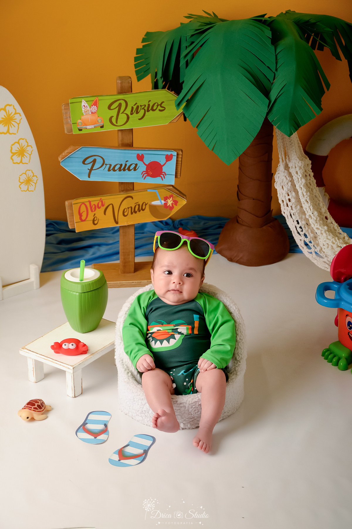 verão2026 - cenário de praia - acompanhamento mesal - drica studio - xinguara - pará - bebê usando roupinha de praia verde - sentado numa cadeirinha branca - com oculos de sol na cabeça - 