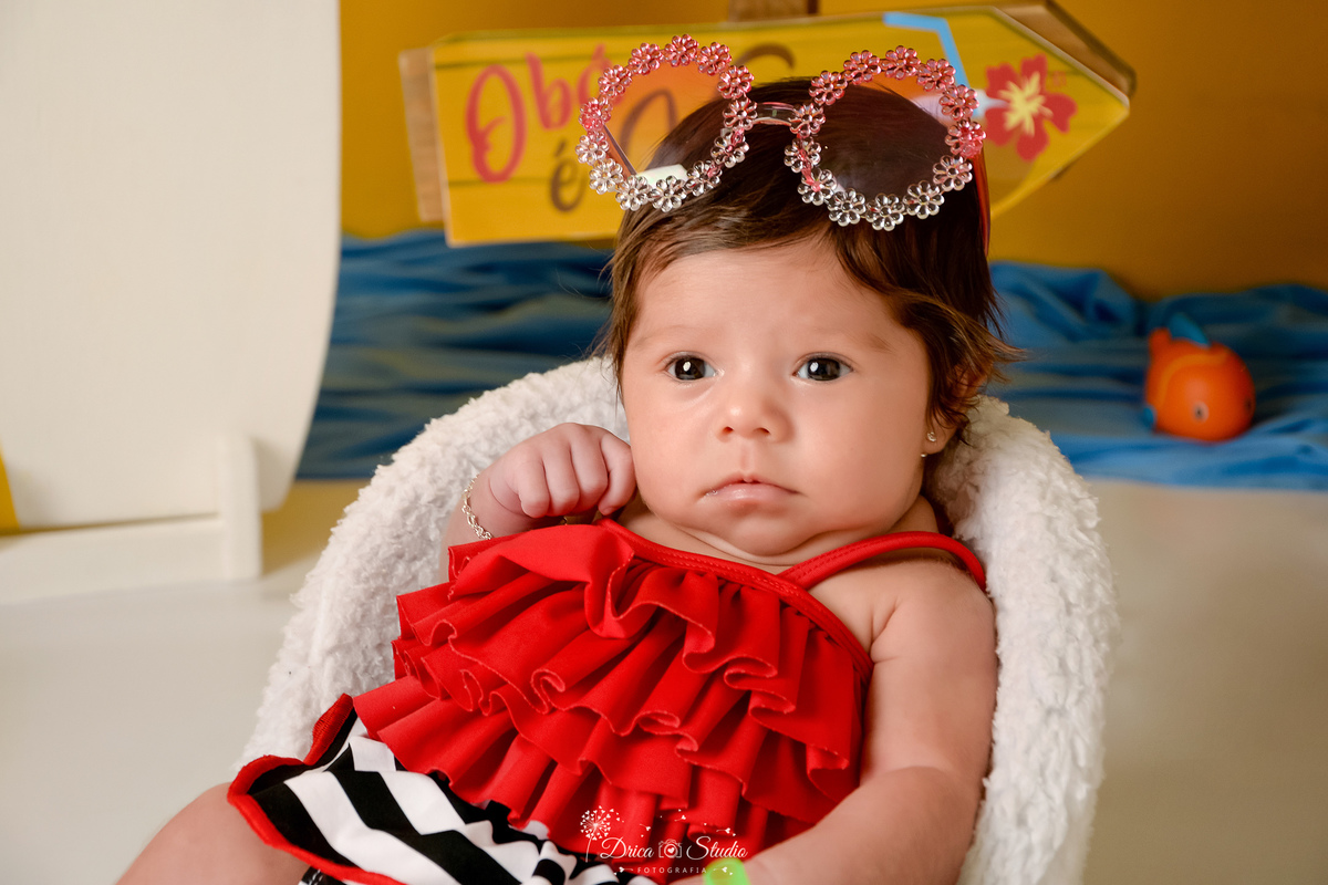 verão2026 - cenário de praia - acompanhamento mesal - drica studio - xinguara - pará - bebe usando roupa de praia na cor vermelha - com oculos de sol na cabeça - sentada em uma cadeirinha branca 