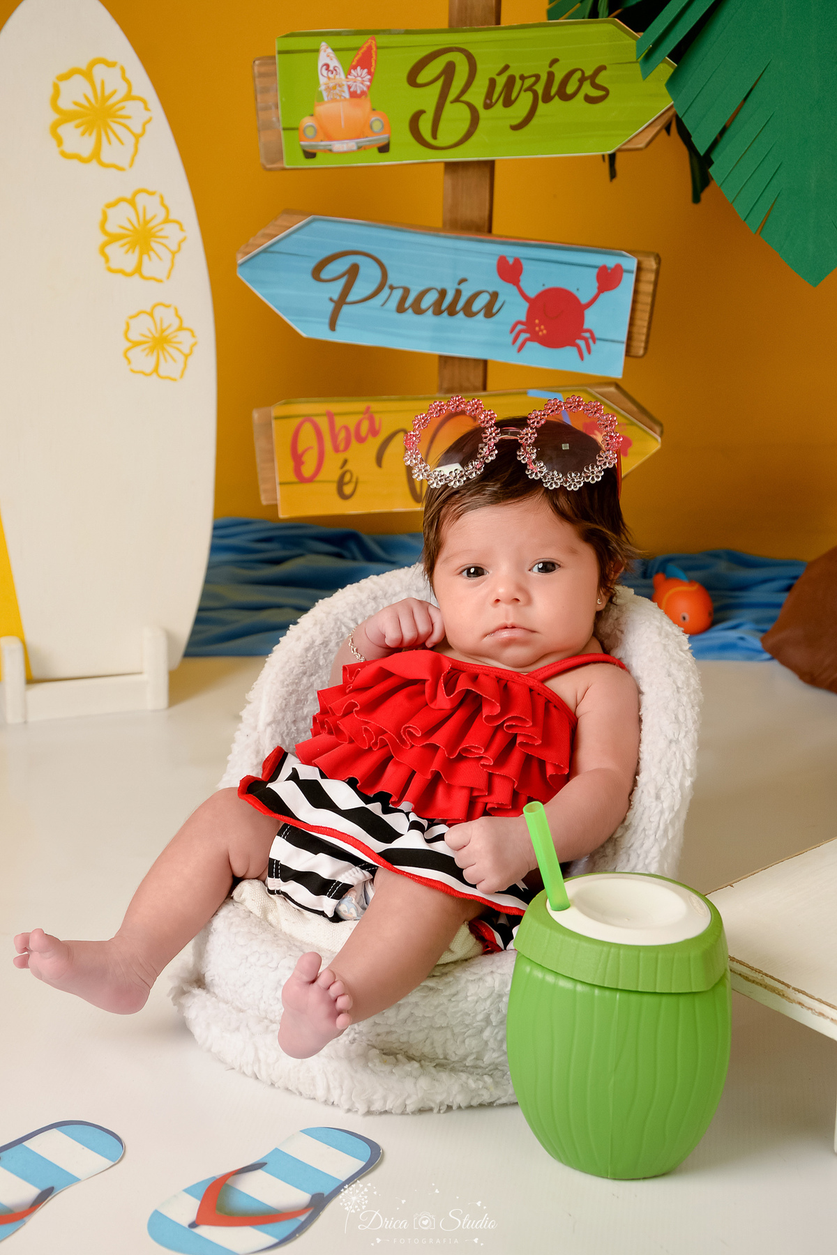 verão2026 - cenário de praia - acompanhamento mesal - drica studio - xinguara - pará  - bebe usando roupa de praia vermelha - sentada em uma cadeirinha branca - com oculos de sol na cabeça 