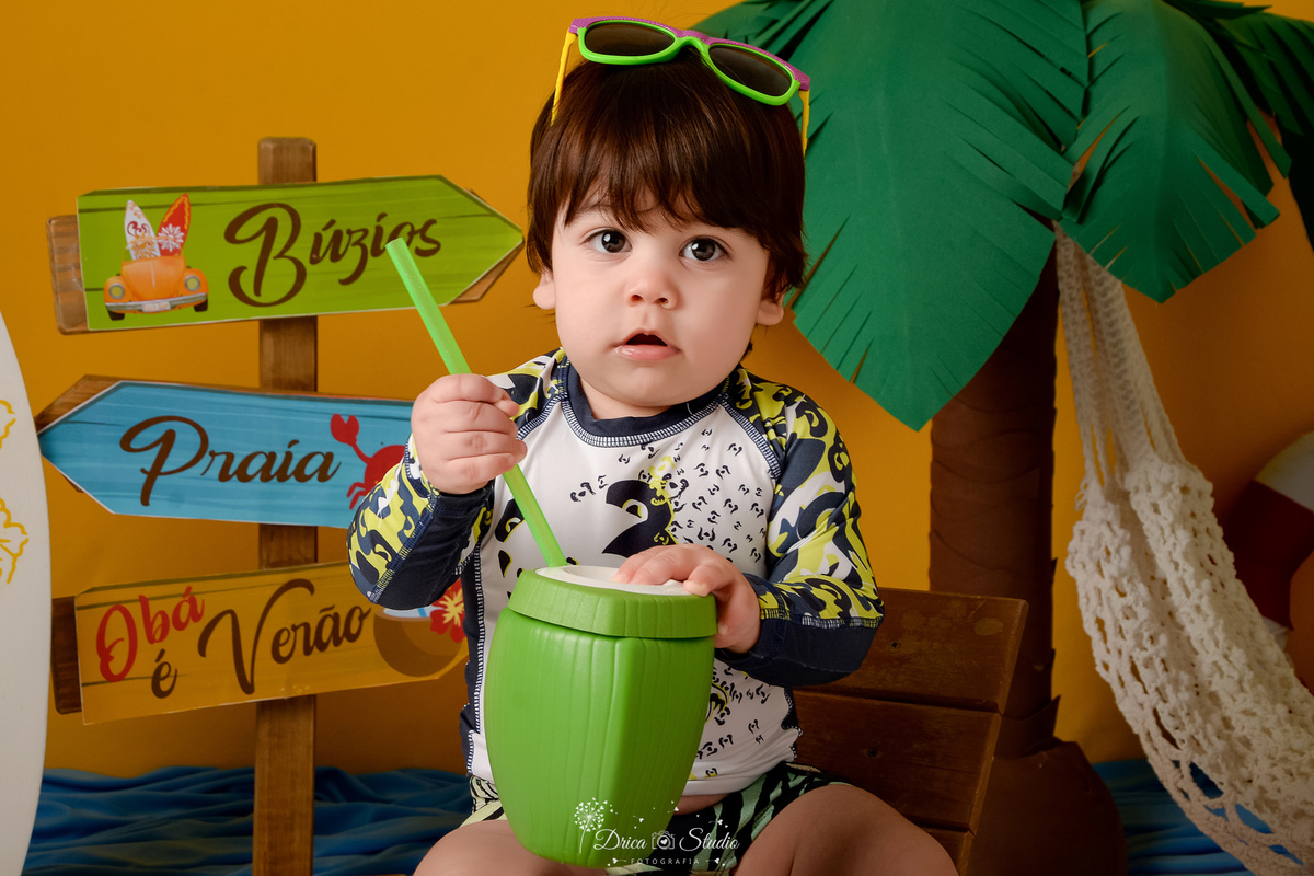 verão2026 - cenário de praia - acompanhamento mesal - drica studio - xinguara - pará - bebe com oculos de sol na cabeça - usando roupa de banho - sentado em uma cadeira de praia - coqueiro verde e marrom ao fundo - rede branca 