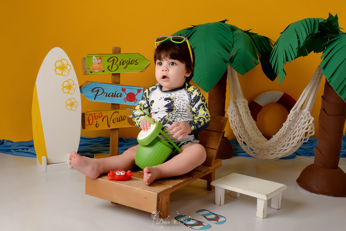 verão2026 - cenário de praia - acompanhamento mesal - drica studio - xinguara - pará - bebe sentado em uma cadeira de praia com oculos de sol na cabeça - usando roupa de banho - coqueiros ao fundo - rede branca - fundo amarelo 