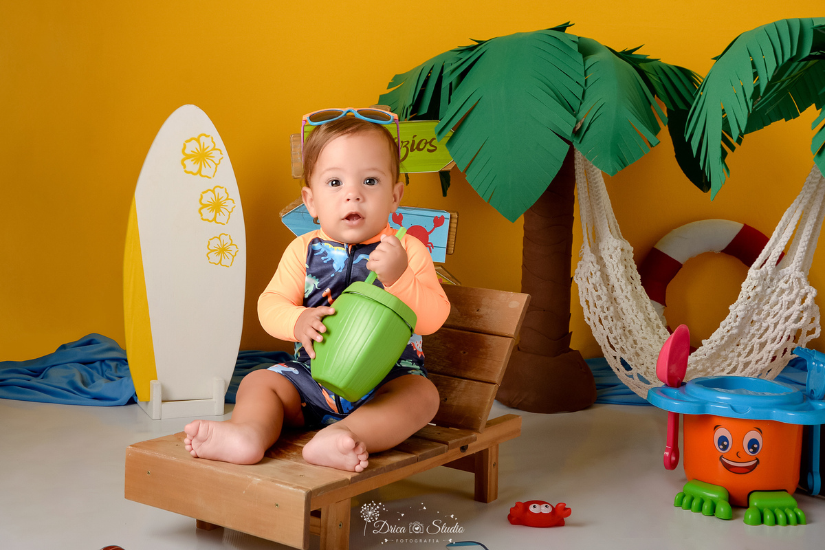 verão2026 - cenário de praia - acompanhamento mesal - drica studio - xinguara - pará bebê sentado em uma cadeira de praia - segurando um copinho em formado de coco - fundo amrelo - com coqueiro marrom  e verde atras - e uma prancha de surfe ao lado 