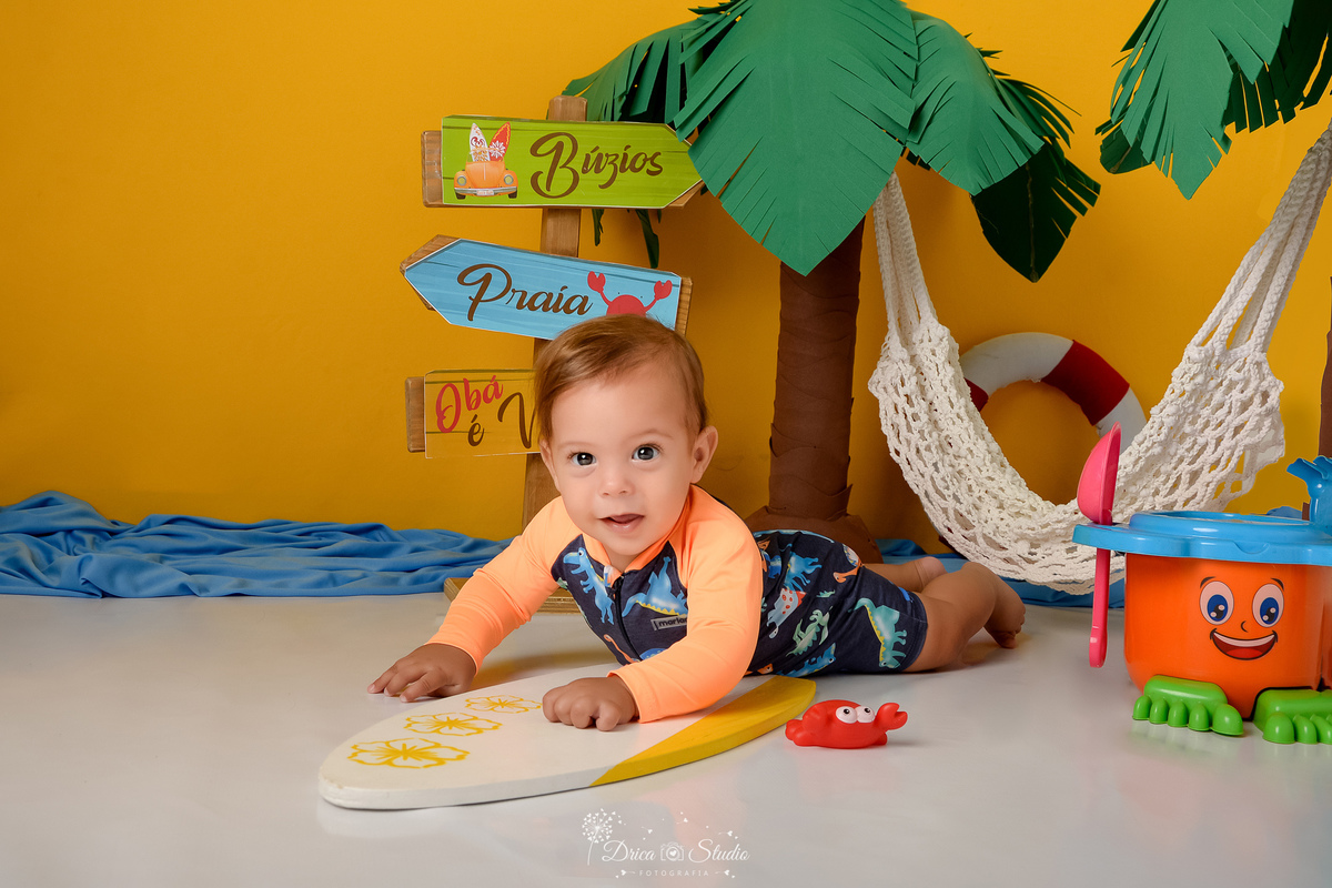 verão2026 - cenário de praia - acompanhamento mesal - drica studio - xinguara - pará - bebe deitado sobre prancha de surfe - fundo amarelo - coqueiro verde com marrom - rede branca - bebe usando roupa de praia 