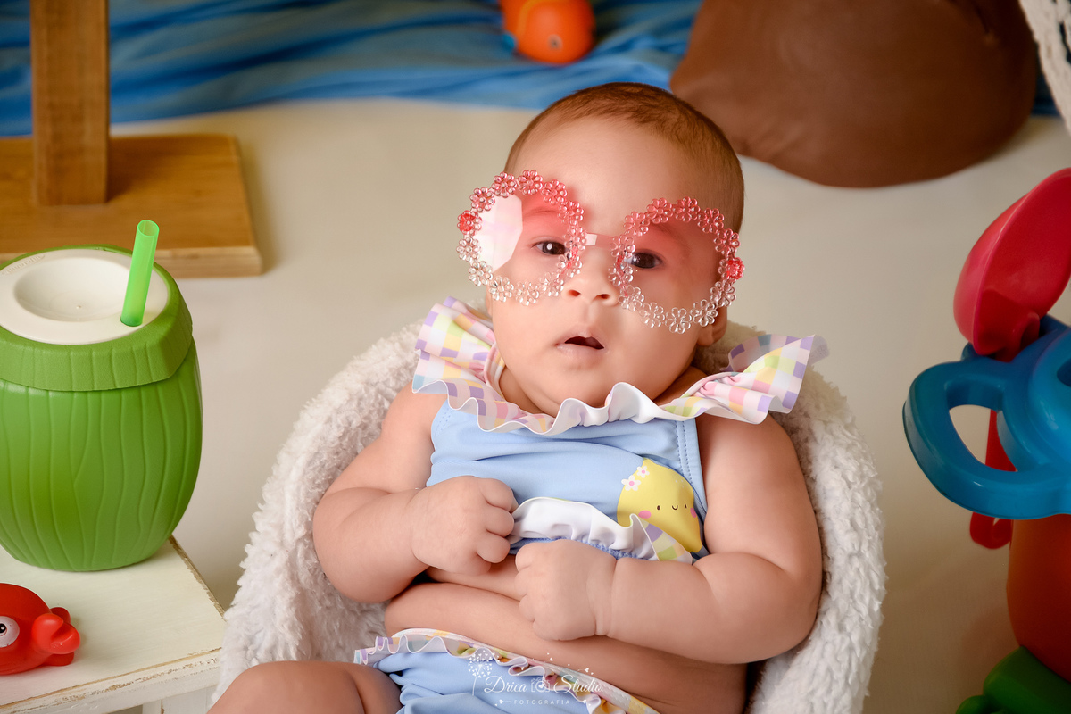 verão2026 - cenário de praia - acompanhamento mesal - drica studio - xinguara - pará bebe sentada em uma cadeirinha branca - usando roupinha de banho - com oculos de sol - ao lado um copo em formato de coco 