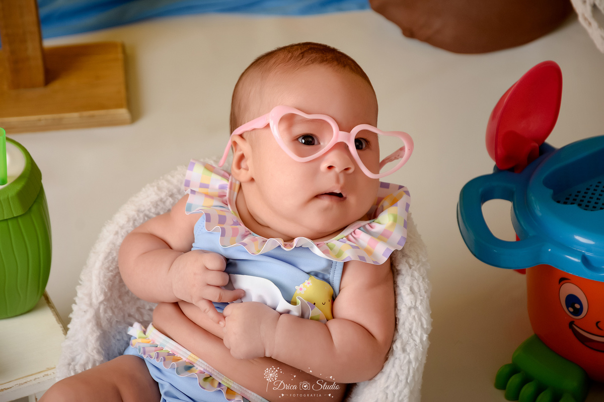 verão2026 - cenário de praia - acompanhamento mesal - drica studio - xinguara - pará - bebe sentada em uma cadeirinha branca - usando roupa de banho - com oculos de coração 