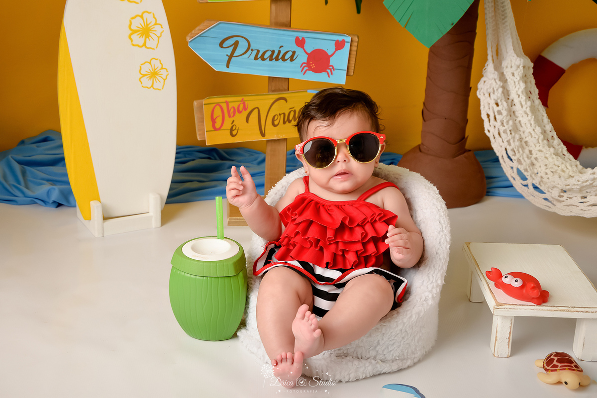 verão2026 - cenário de praia - acompanhamento mesal - drica studio - xinguara - pará - bebe sendata em cadeirinha branca - usando roupa vermelha - ao lado de um copo de coco - fundo amarelo - prancha de surfe branca com dethalhes amarelos 