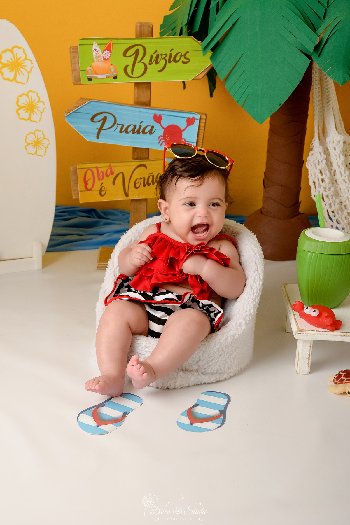 verão2026 - cenário de praia - acompanhamento mesal - drica studio - xinguara - pará - bebe sentada em cadeira de praia branca com roupa de praia vermelha - oculos de sol na cabeça - sorrindo para a foto 