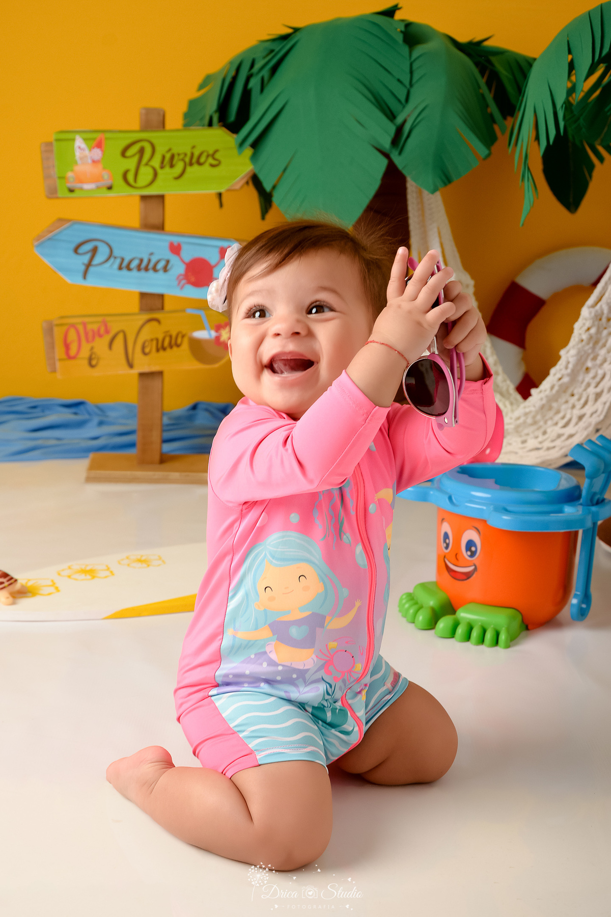 verão2026 - cenário de praia - acompanhamento mesal - drica studio - xinguara - pará - bebê usando roupinha de banho rosa - com oculos de sol rosa na mão ao fundo um baldinho de areia e uma prancha de surfe 