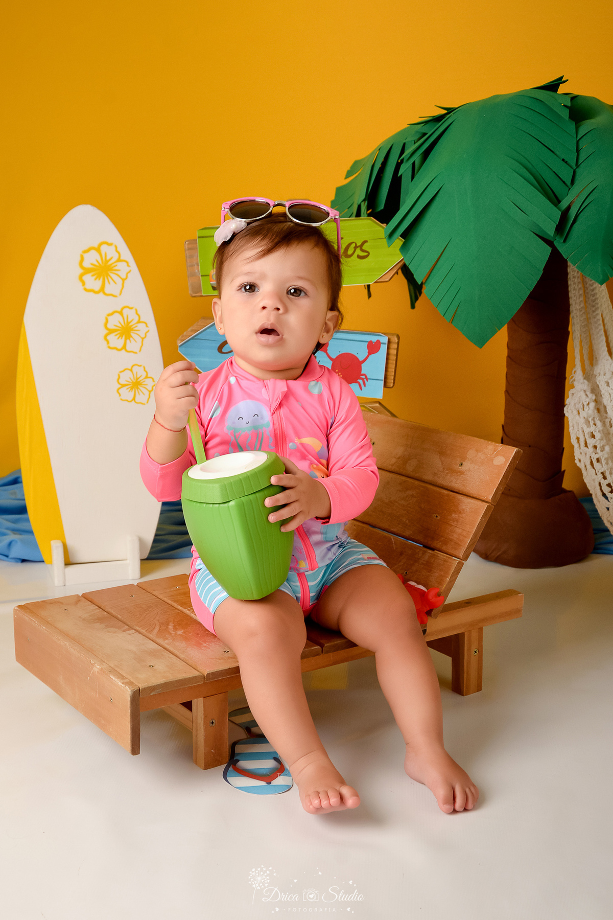 verão2026 - cenário de praia - acompanhamento mesal - drica studio - xinguara - pará - bebe sentada em cadeira de praia - com copo de coco na mão - ao fundo uma prancha de surfe branca com dethalhes amarelos 