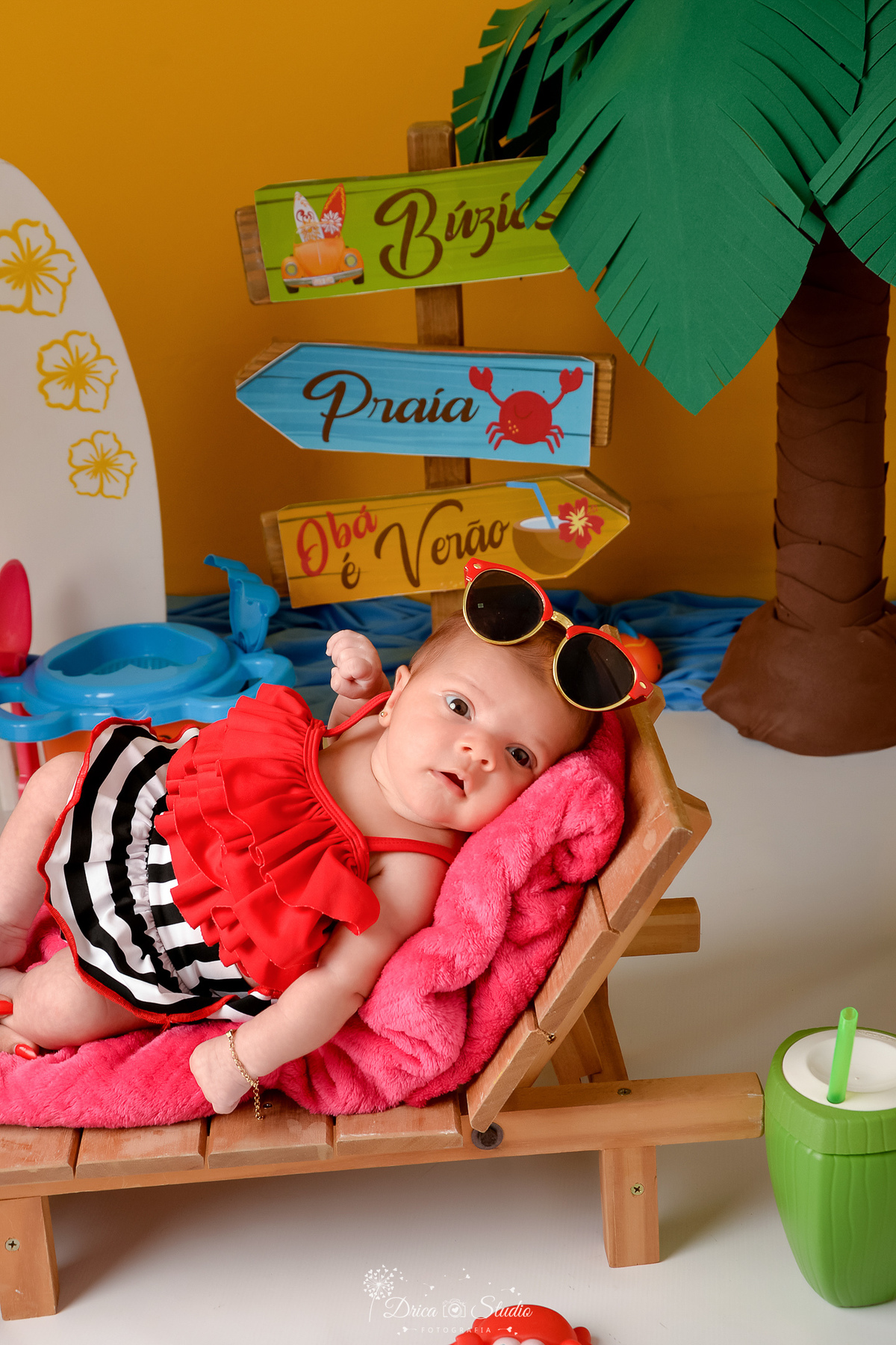 verão2026 - cenário de praia - acompanhamento mesal - drica studio - xinguara - pará - bebe deitada em cadeira de praia - usando roupa de banho vermelha com preto e branco - com oculos de sol na cabeça 
