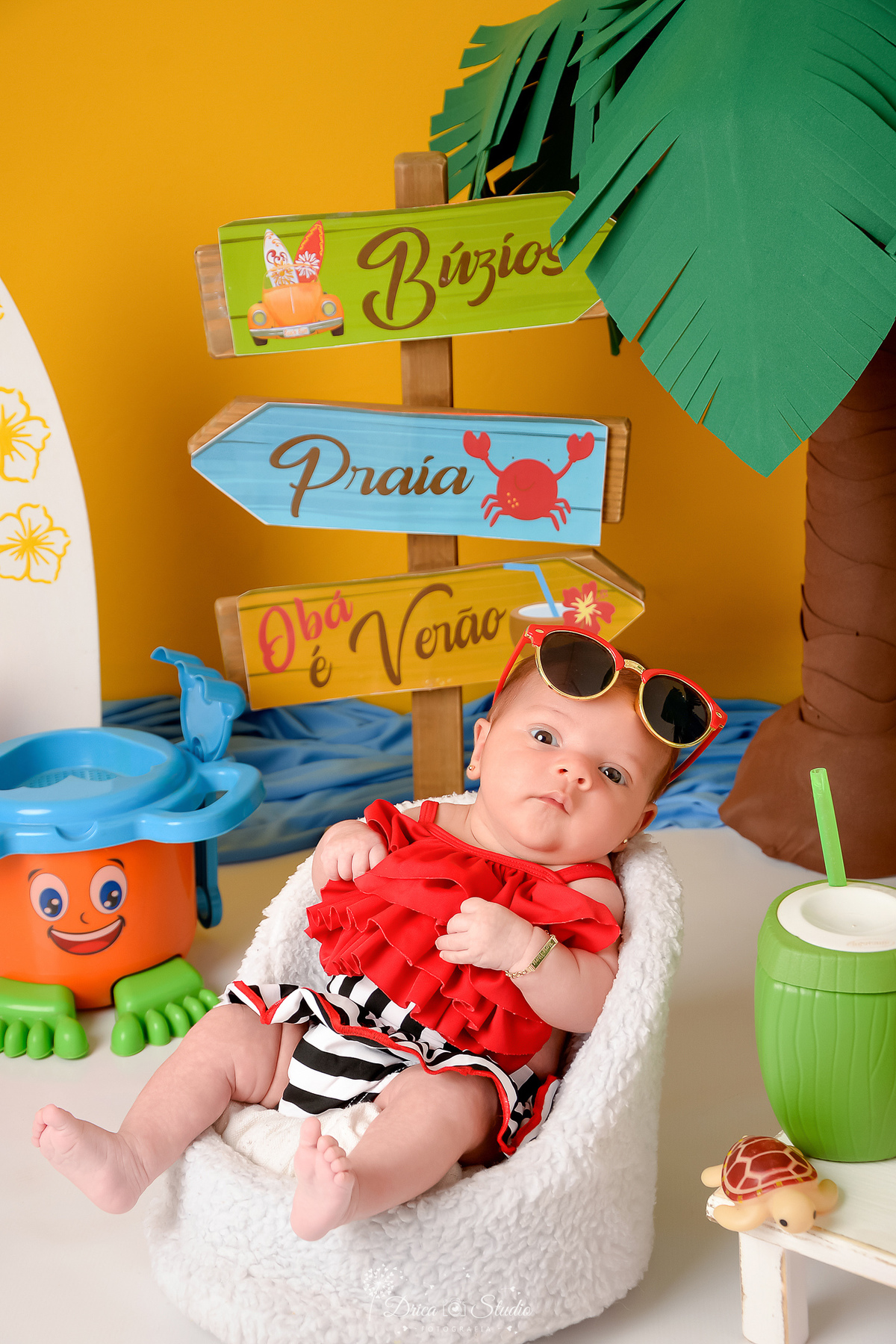 verão2026 - cenário de praia - acompanhamento mesal - drica studio - xinguara - pará - bebe sentada em uma cadeirinha branca - com roupa de banho vermelha - com oculos de sol na cabeça 