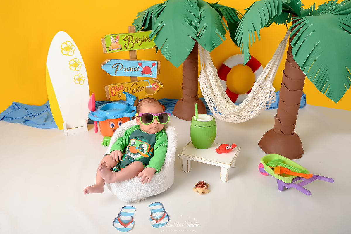 verão2026 - cenário de praia - acompanhamento mesal - drica studio - xinguara - pará - bebe sentado em cenário de praia - usando roupa de banho - com oculos de sol - ao fundo 2 coqueiros - cim uma rede branca - ao lado de uma prancha de surf 