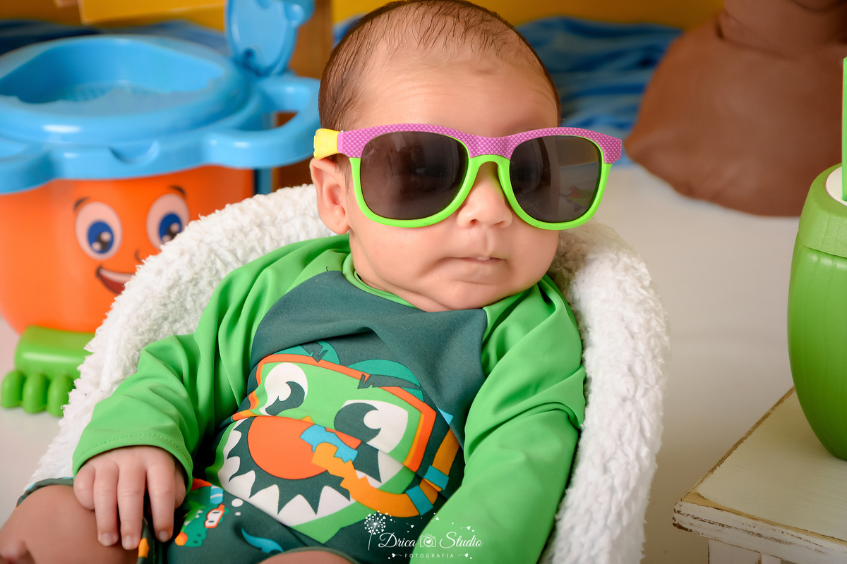 verão2026 - cenário de praia - acompanhamento mesal - drica studio - xinguara - pará - bebe usando roupa de banho verde - com oculos de sol - sentado em cadeirinha branca 