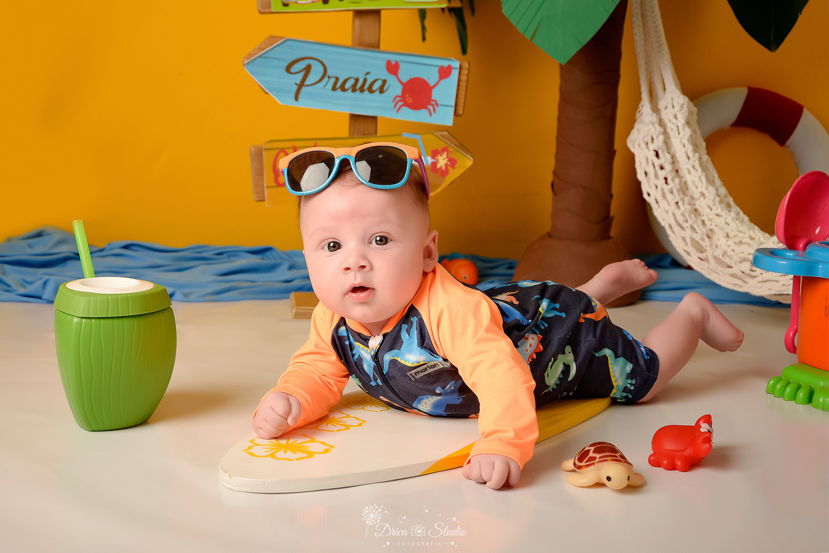 verão2026 - cenário de praia - acompanhamento mesal - drica studio - xinguara - pará - bebe deitado em prancha de surfe - com oculos de sol na cabeça - usando roupa de banho - ao lado de um copo de coco - 