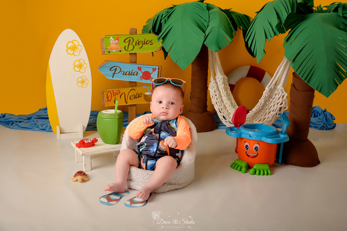 verão2026 - cenário de praia - acompanhamento mesal - drica studio - xinguara - pará - bebe sentado em cadeira branca - com roupa de banho - com osculos de sol na cabeça - fundo amarelo - com coqueiros e uma rede branca 