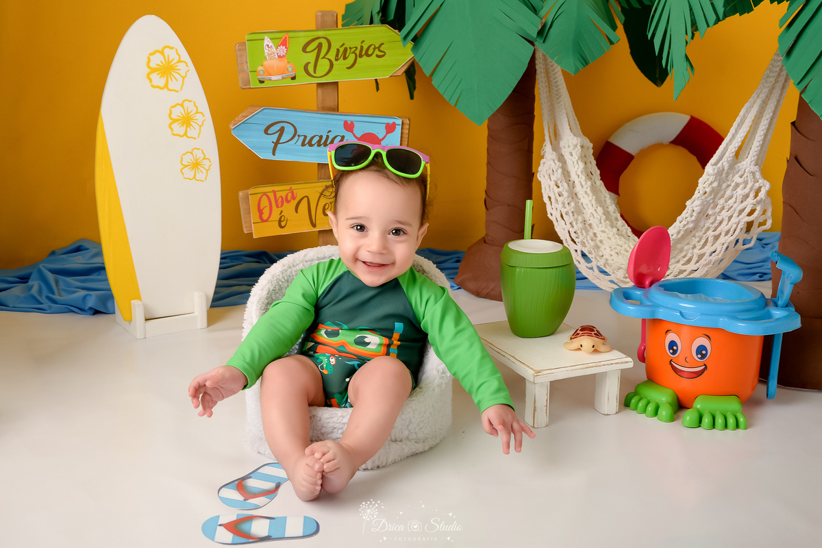 verão2026 - cenário de praia - acompanhamento mesal - drica studio - xinguara - pará - bebe sentado em uma cadeira branca - com roupa de praia verde - com oculos de sol na cabeça - ao lado um prancha de surfe 