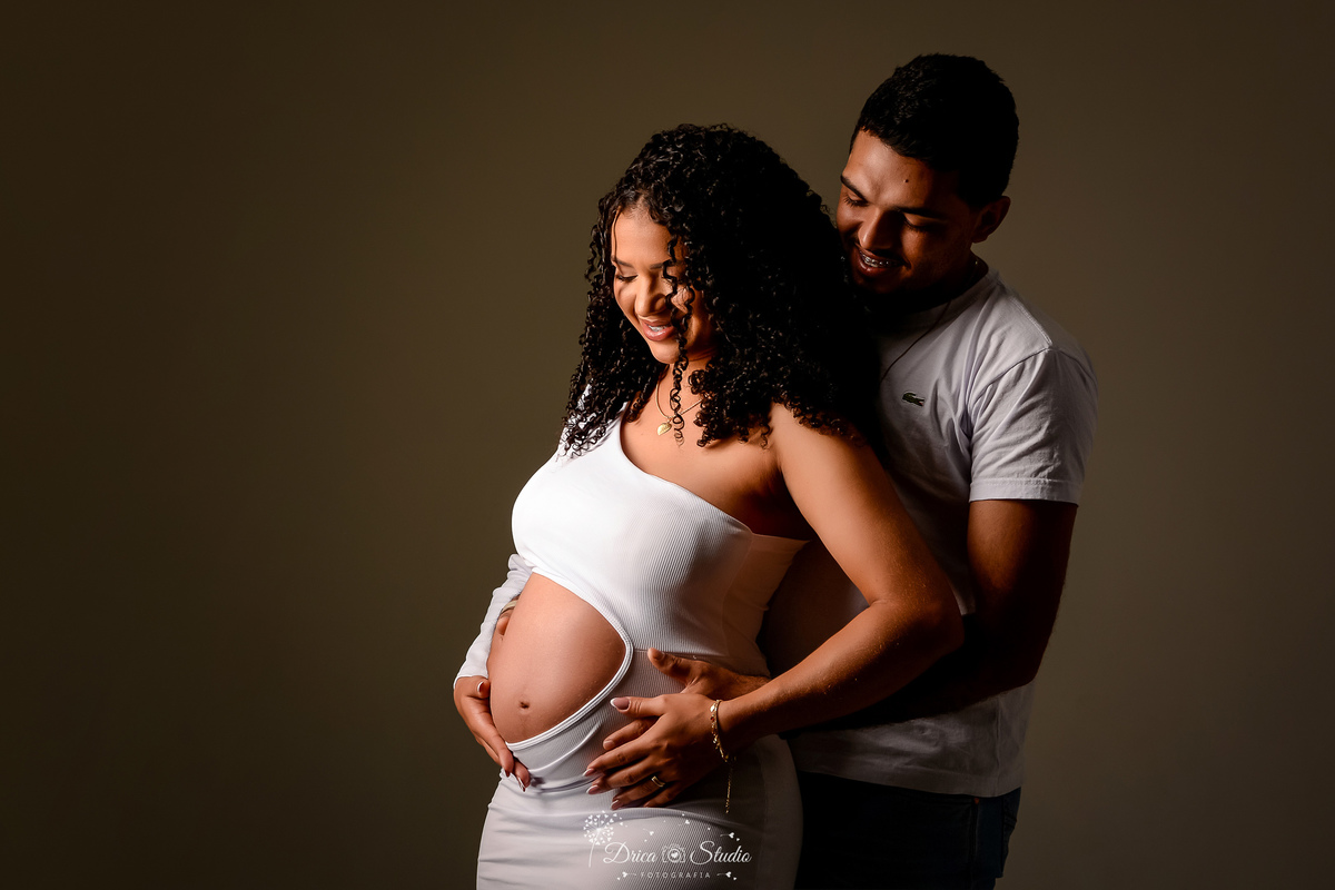 drica studio - ensaio gestante - primeiro filho - fotos em estudio - xinguara - pará - mãe usando vestido branco que mostra a barriga - pai com blusa branca e calça jeans - fundo escuro 