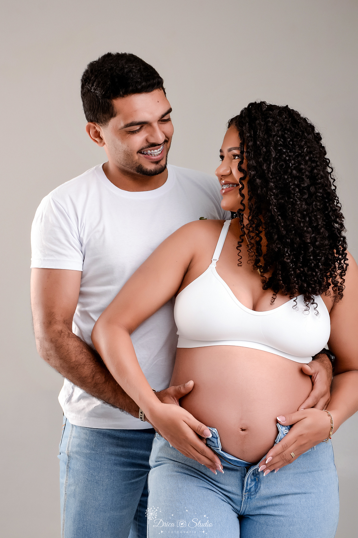 drica studio - ensaio gestante - primeiro filho - fotos em estudio - xinguara - pará - mãe usando top branco com calça jeans - pai usando camisa branca  - se ohando com as mãos na barriga 