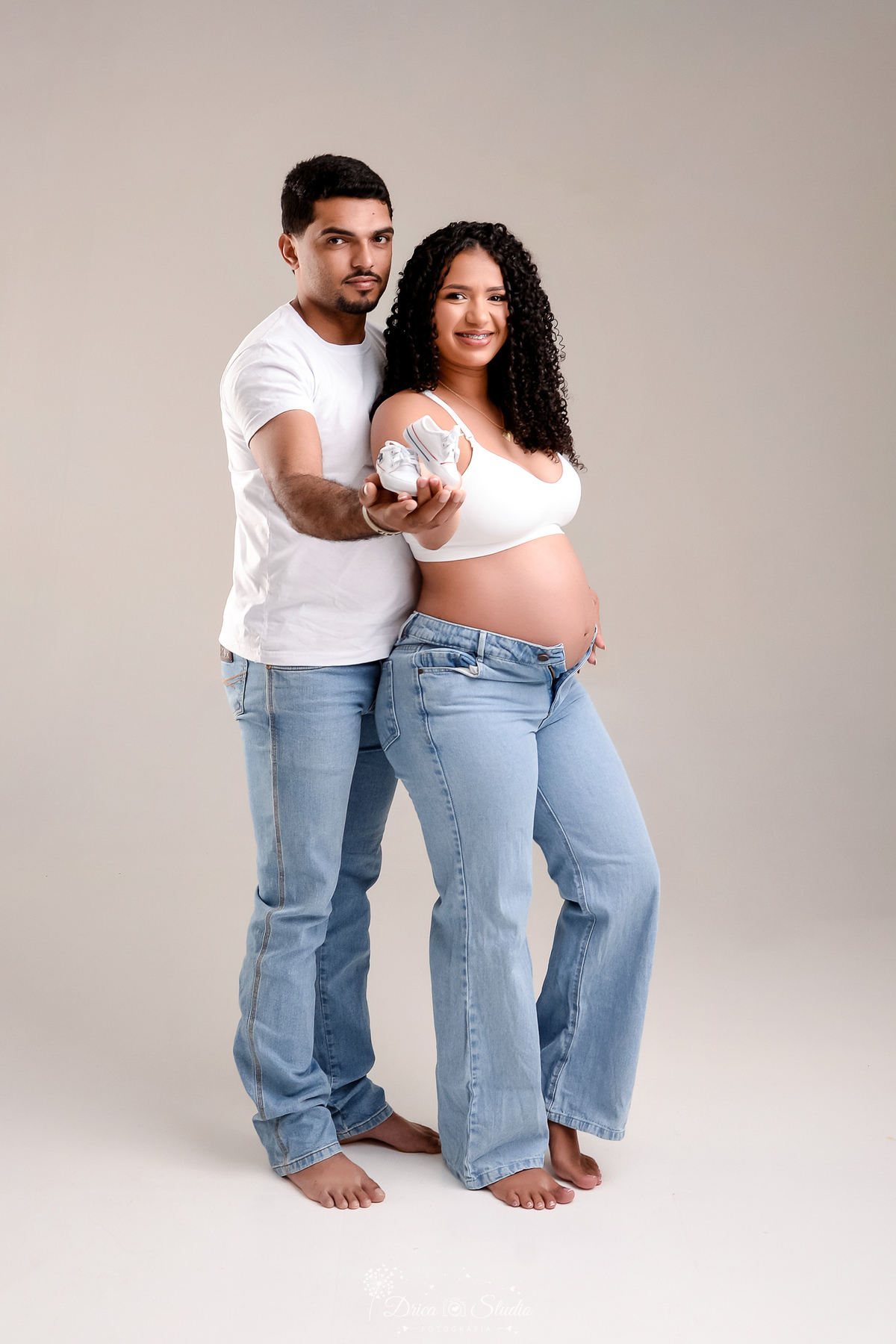 drica studio - ensaio gestante - primeiro filho - fotos em estudio - xinguara - pará - pai usando camisa branca com calça jeans - mãe usando top branco com calça jeans - segurando sapatinho do bebê  - mãe com a mão na barriga 