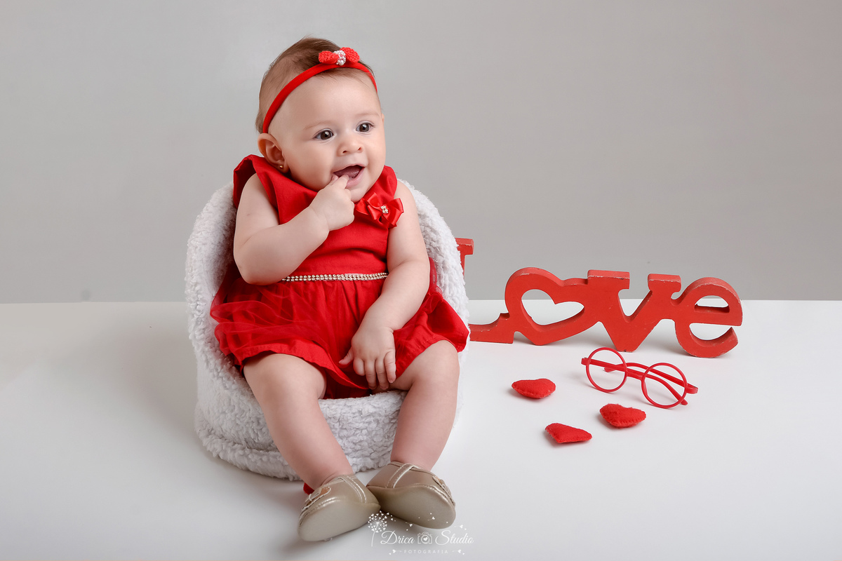 drica studio - acompanhamento mesal - 4 meses - xinguara - pará - foto em studio - bebe sentada em cadeirinha branca -  usando vestido vermelho com laço vermelho - ao lado corações e ummini oculos - enfeite love 