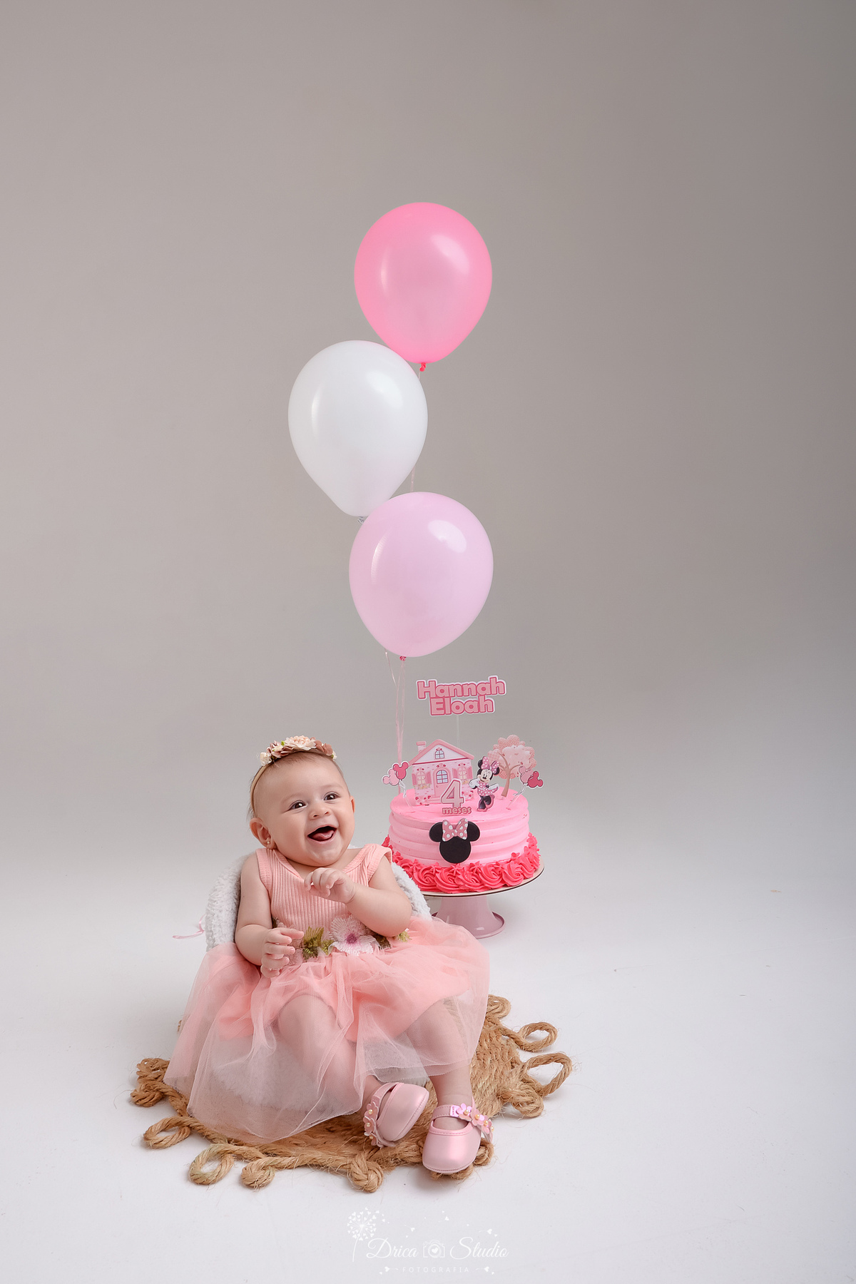 drica studio - acompanhamento mesal - 4 meses - xinguara - pará -foto em studio - balão rosa e branco - bolo da minie - bebe sentado em uma baquinho branco - usando vestido e laço rosa - sorrindo para a foto 